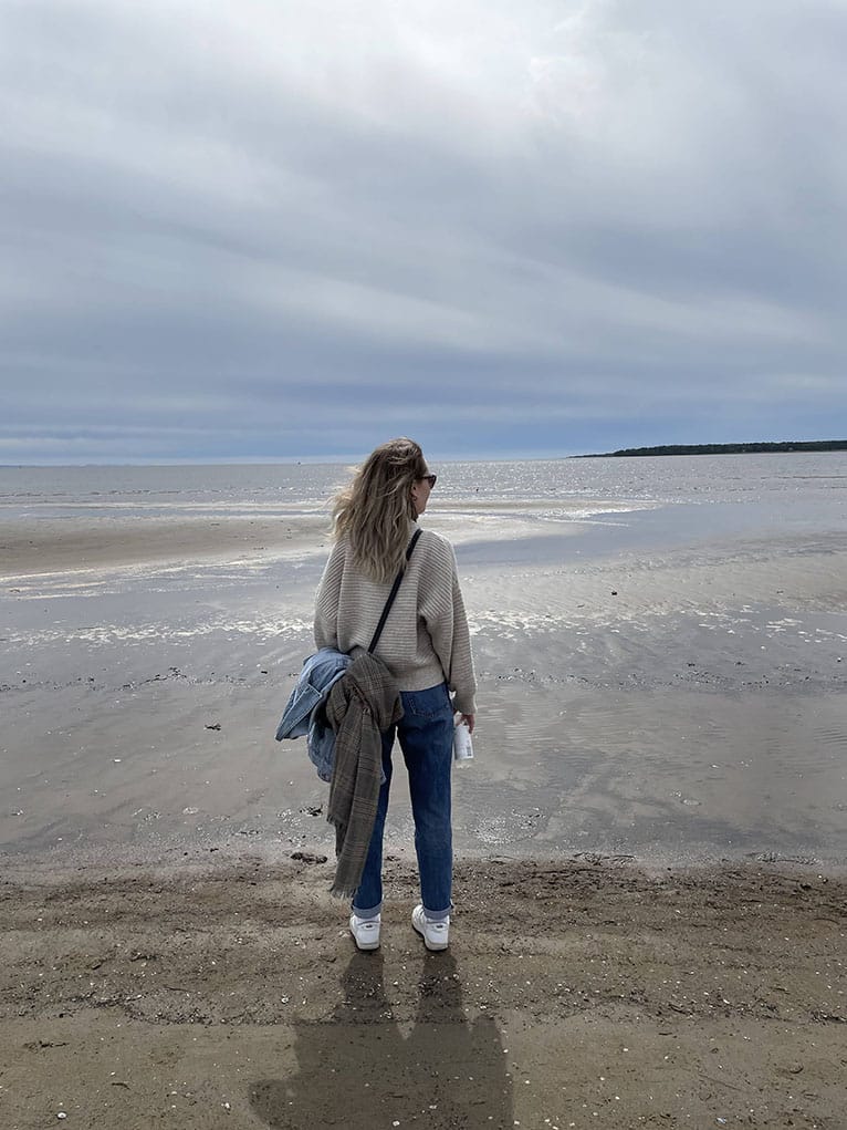 Me, a young woman with shoulder-length blonde hair, wearing sunglasses, a beige hoodie and jeans. I am standing at the beach and looking into the distance.